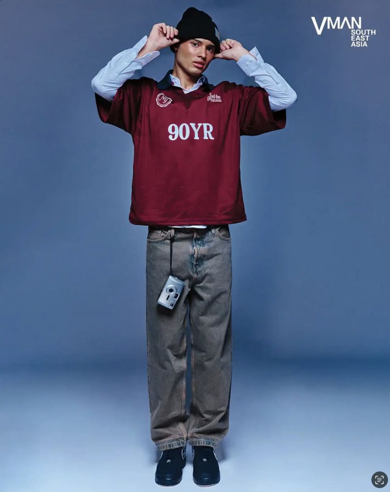 Full-length shot of a young adult male in a maroon t-shirt, baggy jeans, beanie, and black sneakers posing in a blue studio.
