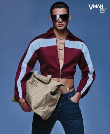 Male fashion model in a cropped jacket, jeans, sunglasses, and jewelry, holding a canvas bag in a blue photo studio.