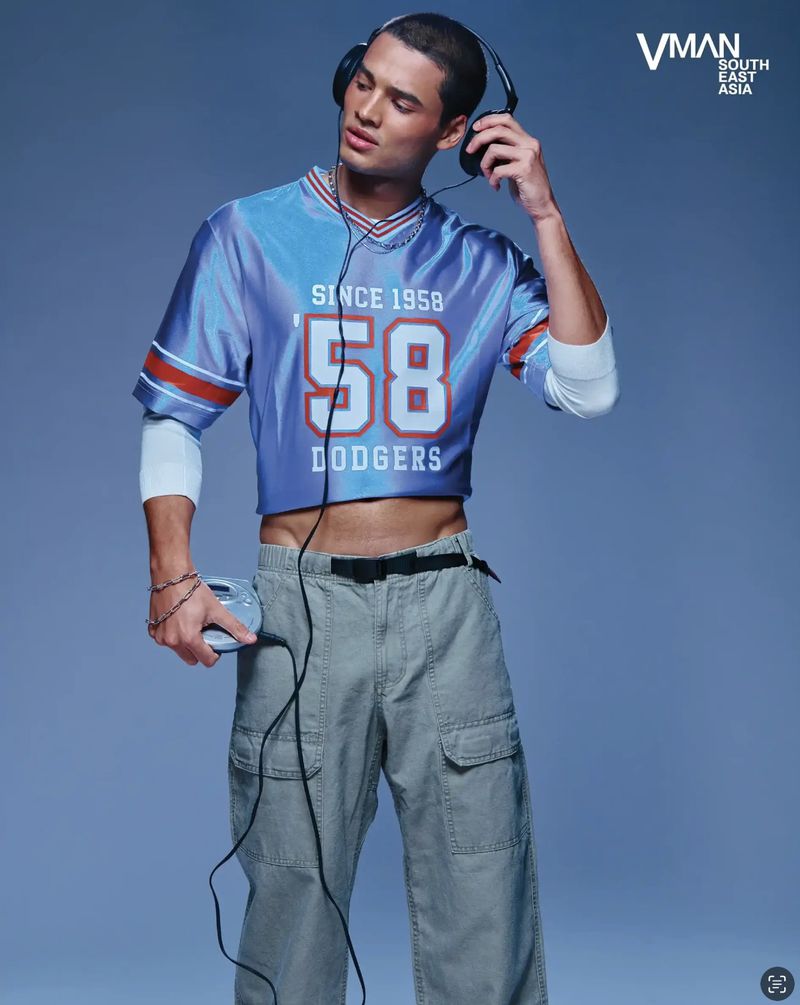 Fashion shot of a young adult male model wearing headphones, a cropped blue t-shirt, and cargo pants against a blue background.