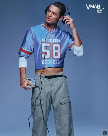 Fashion shot of a young adult male model wearing headphones, a cropped blue t-shirt, and cargo pants against a blue background.
