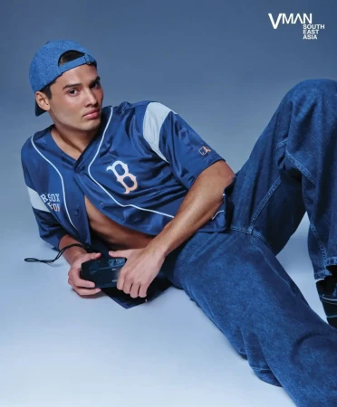 Man in a blue baseball jersey and denim jeans, poses with a vintage camera in a studio setting, lit with soft, diffused lighting.