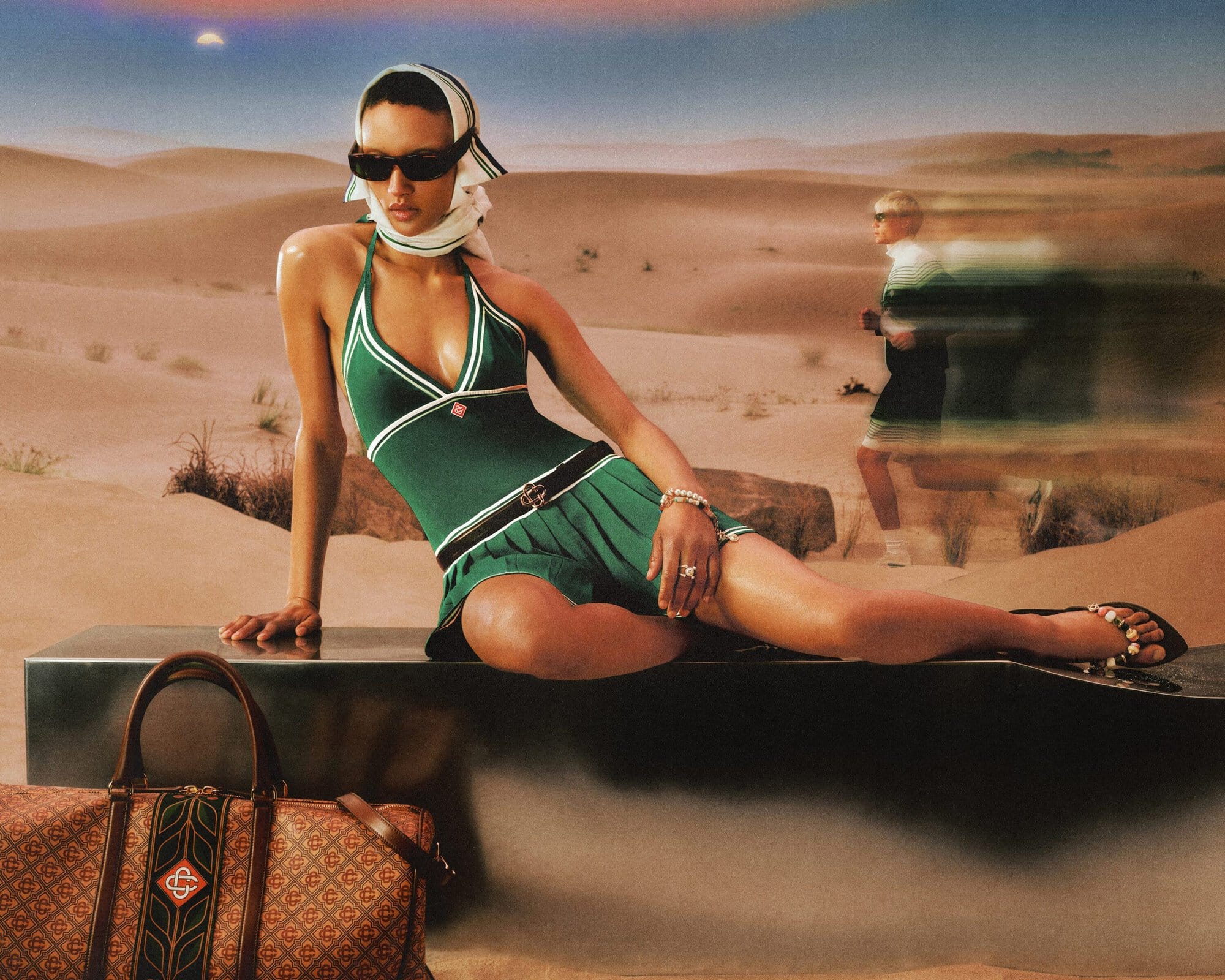 A woman in a green swimsuit and headscarf poses with a patterned bag in a desert landscape on a sunny day. Fashion photoshoot.