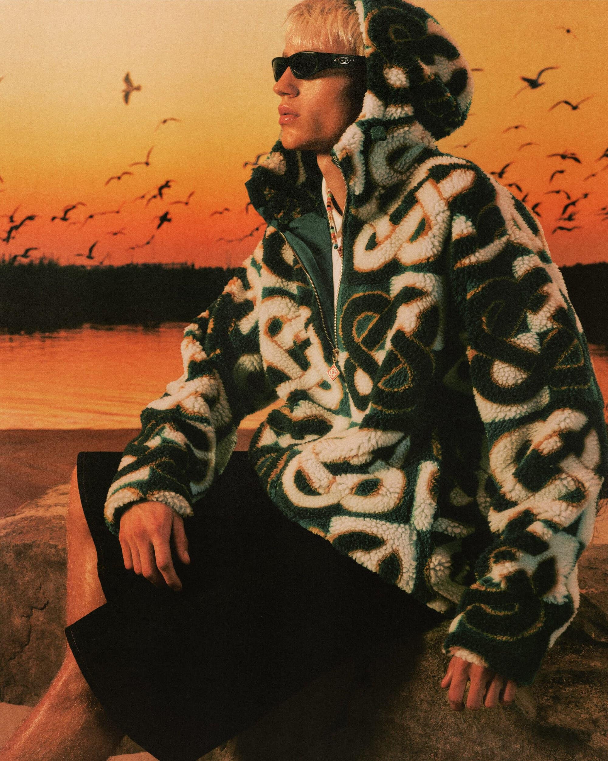 Model wearing a patterned green fleece hoodie and dark shorts seated outdoors against a sunset sky filled with birds.