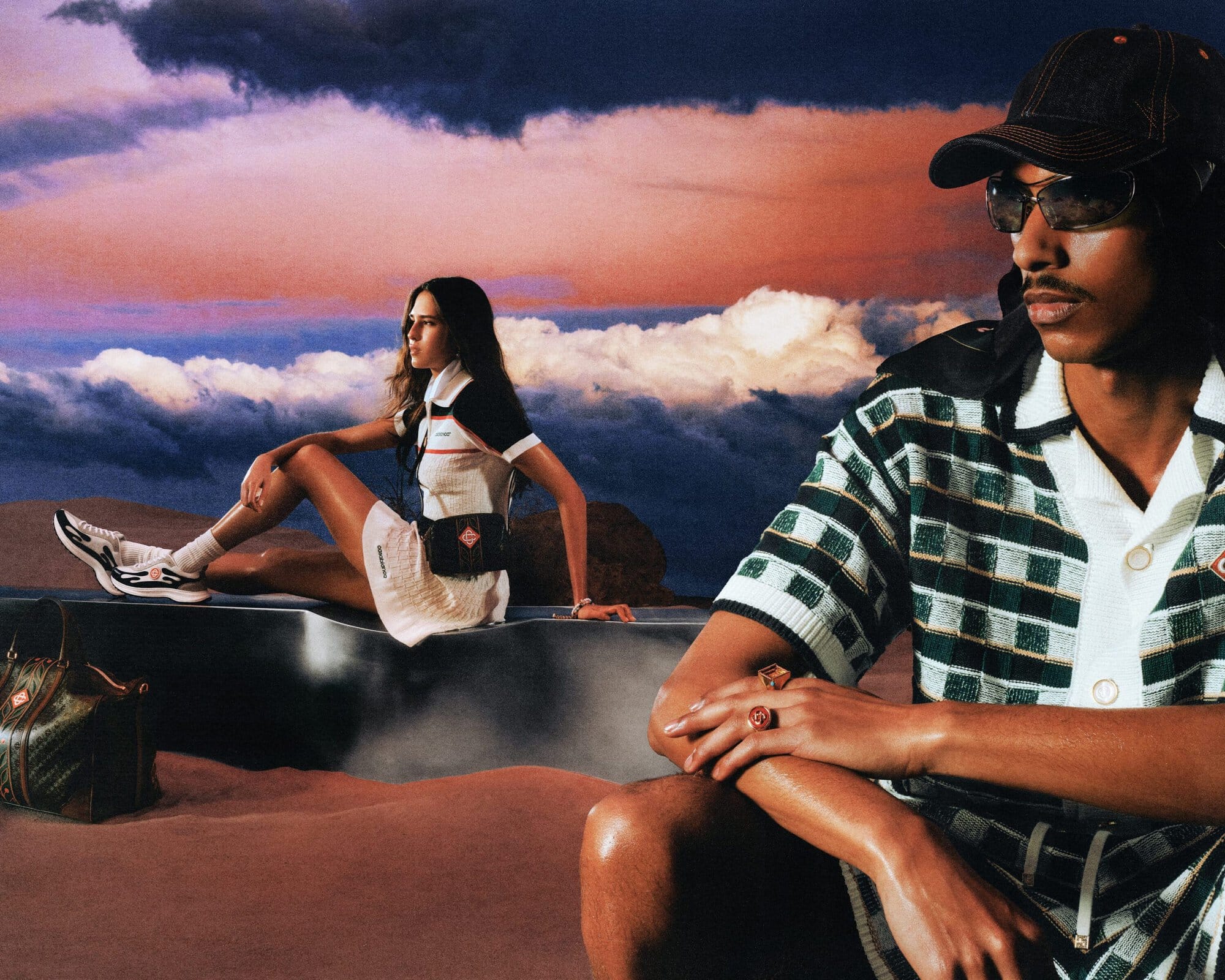 Man in patterned knit polo and cap foregrounded against a dramatic sunset sky with a seated woman in white