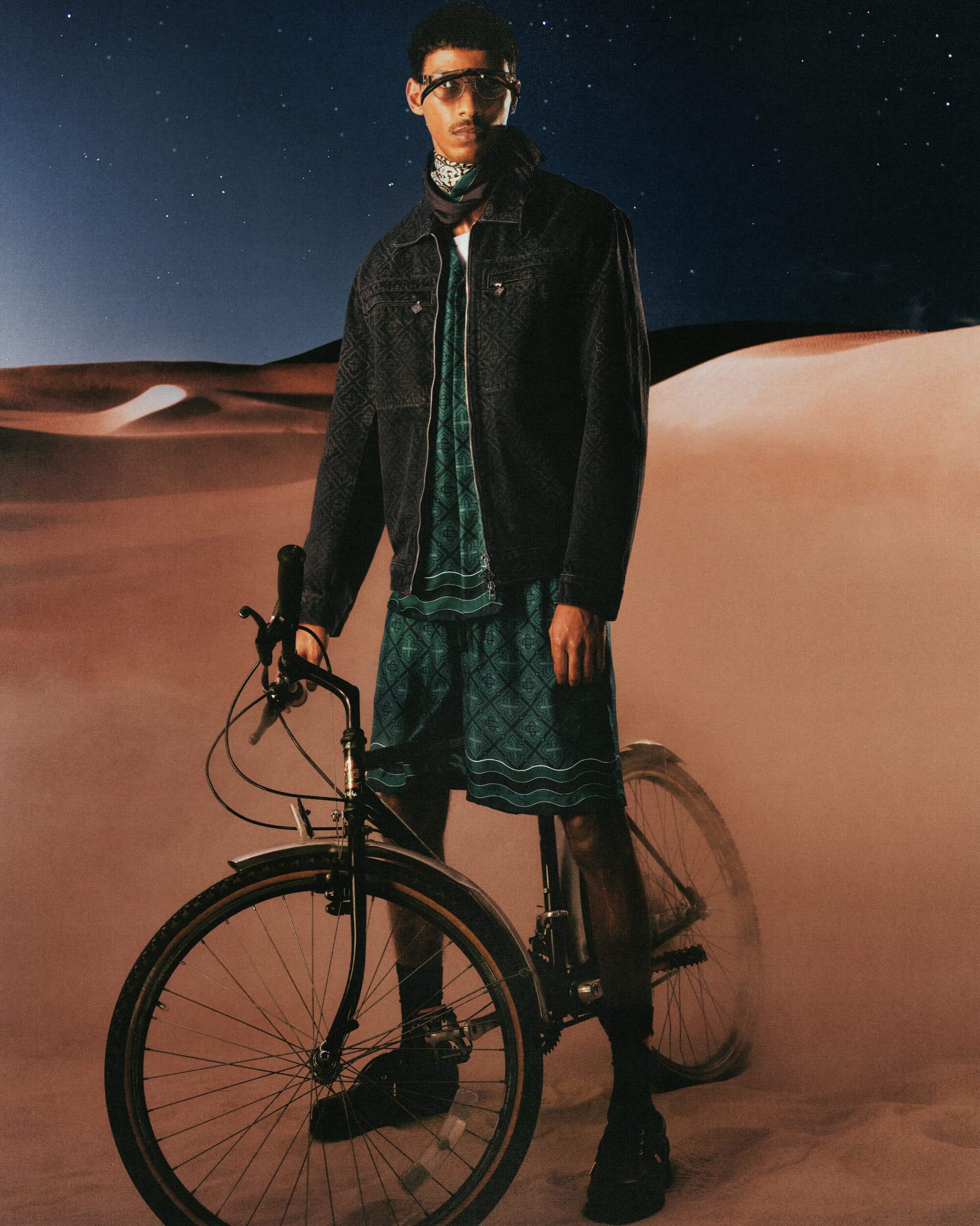 A man wearing a black jacket and patterned green shorts stands outdoors holding a bicycle against a desert landscape at night.