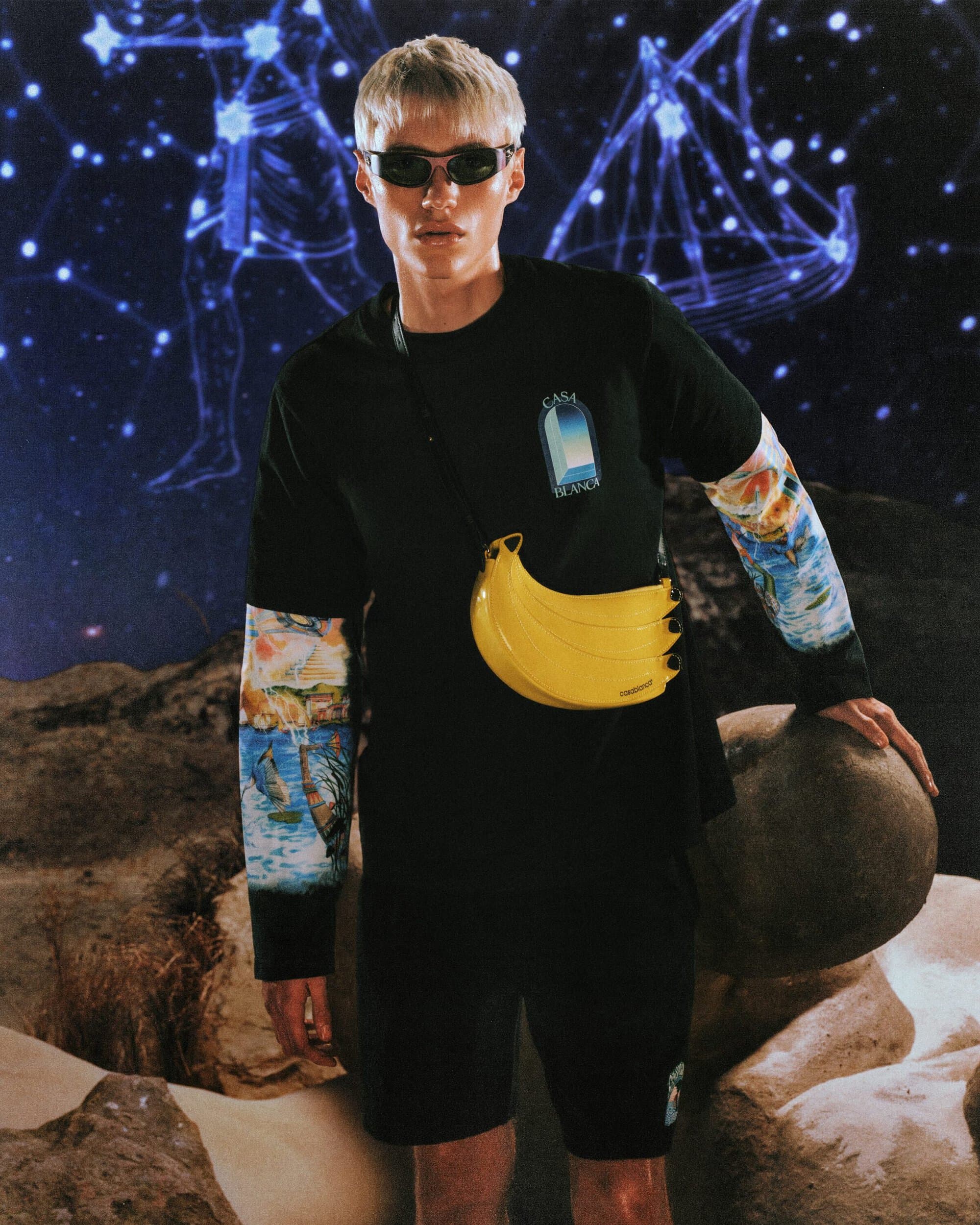 Blonde male model wearing dark sunglasses, black t-shirt with graphic, and yellow banana-shaped crossbody bag against a starry backdrop