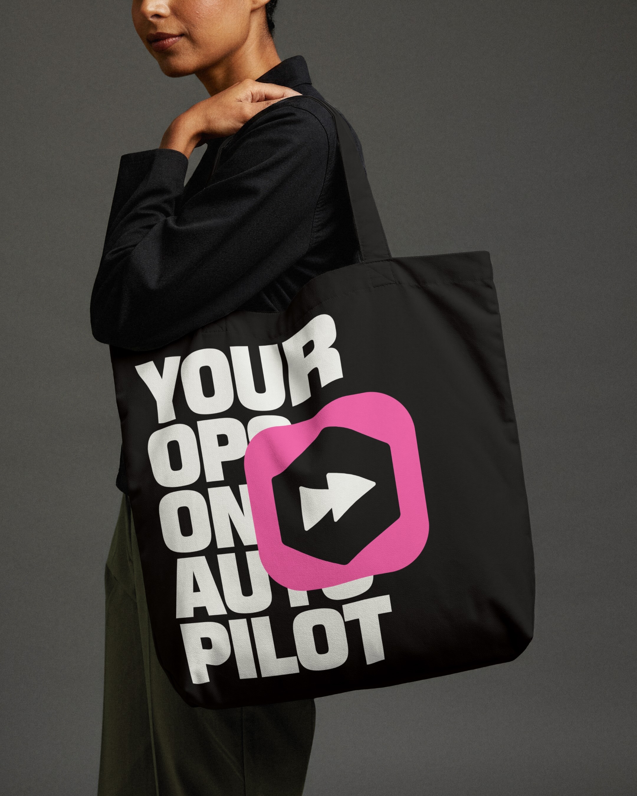 A person holds a black tote bag with white text and a pink graphic design element against a neutral background in a studio shot.