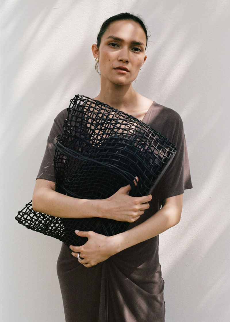 Adult female poses against a white wall, wearing a brown dress and holding a black mesh bag, bathed in soft, diffused natural light.