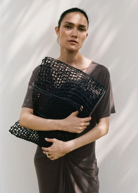 Adult female poses against a white wall, wearing a brown dress and holding a black mesh bag, bathed in soft, diffused natural light.