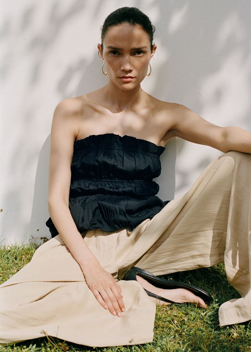 A woman wearing a black tube top and beige pants sits in the grass outdoors in the sunlight. Fashion editorial lifestyle shot.