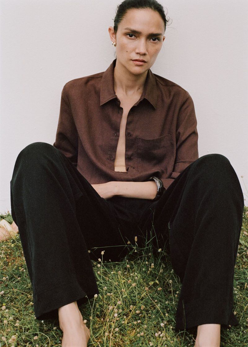 Portrait of a relaxed woman sitting on grass in a brown shirt and black pants, captured in soft, natural light against a plain backdrop.