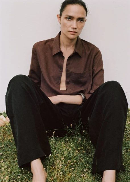 Portrait of a relaxed woman sitting on grass in a brown shirt and black pants, captured in soft, natural light against a plain backdrop.