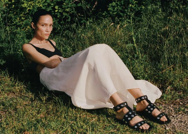 Charlotte Carey reclining outdoors in a black top, flowing white maxi skirt, and studded black flat sandals on grass.