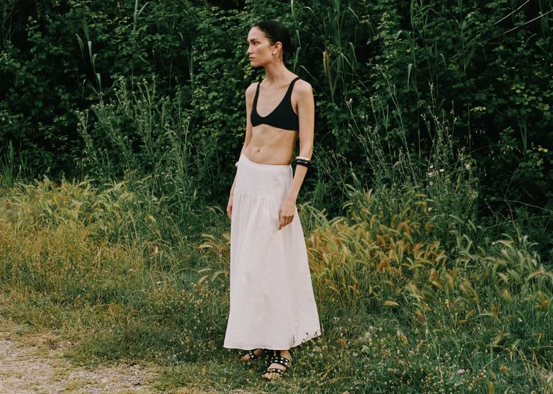 Adult woman in a black top and white skirt standing in a grassy field with trees in the background, outdoors.
