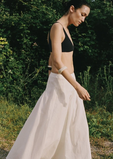 A fashionable woman walks outdoors wearing a black top and white skirt, with lush green foliage in the background under natural light.