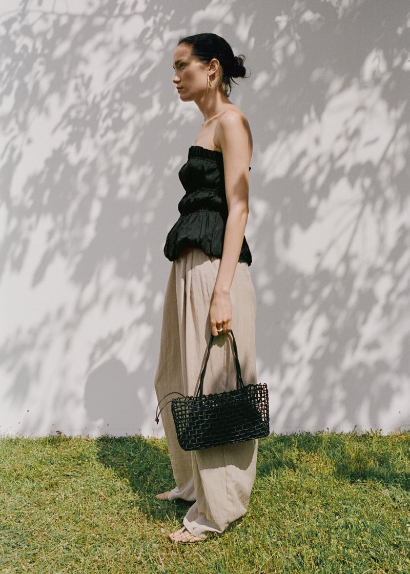 Woman poses outdoors in a black top and khaki pants, holding a black woven handbag, with shadows of leaves on a white wall.