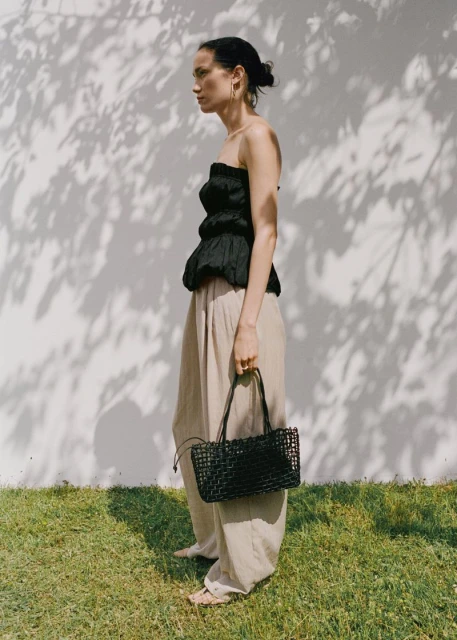 Woman poses outdoors in a black top and khaki pants, holding a black woven handbag, with shadows of leaves on a white wall.