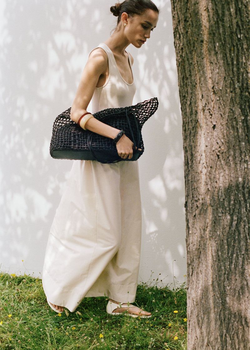 Woman in a neutral dress walks past a tree with a woven handbag. The background is a light wall with a shadow pattern.