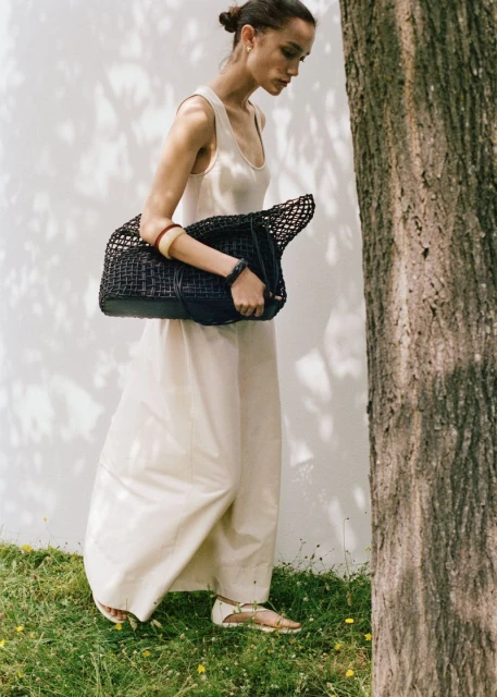 Woman in a neutral dress walks past a tree with a woven handbag. The background is a light wall with a shadow pattern.