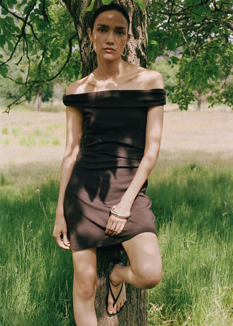 A woman in a short, dark dress leans against a tree in a meadow with long grass, wearing sandals and a bracelet. Sunlight filters through the leaves.