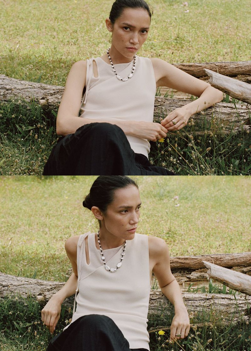Adult female model wearing a necklace and sleeveless shirt sitting on a log outdoors in a grassy field with natural daylight.
