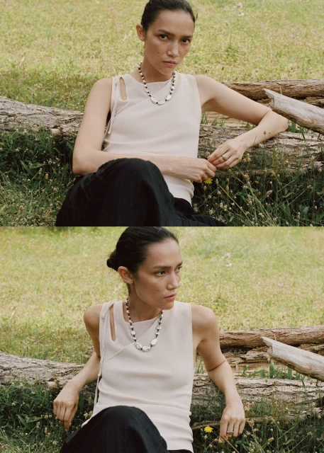 Adult female model wearing a necklace and sleeveless shirt sitting on a log outdoors in a grassy field with natural daylight.