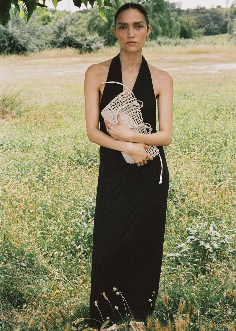 Adult female in a long black dress standing in an outdoor field with trees, holding a white woven bag, with soft natural lighting.
