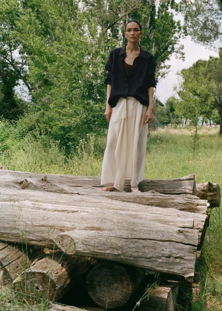 Adult female in a black button-down shirt and white skirt standing on a pile of logs in a grassy, wooded area on a sunny day.