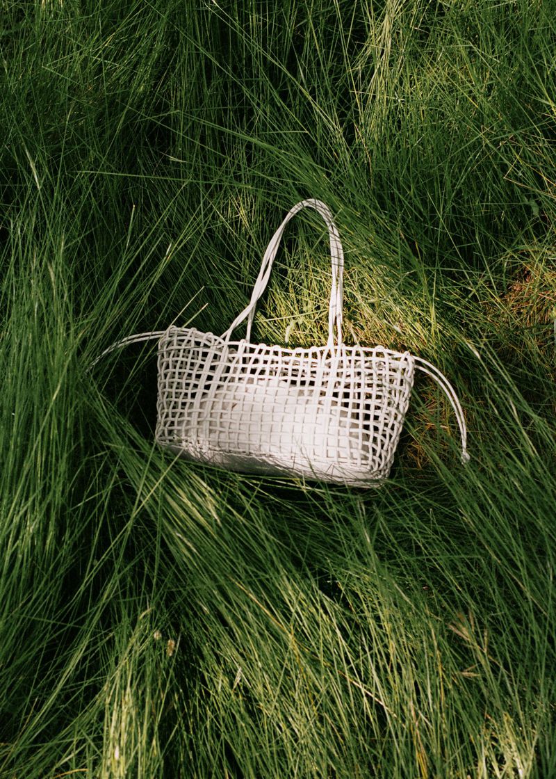 Woven white tote bag with handles sits in a field of long green grass, showcasing a nature-inspired fashion accessory.