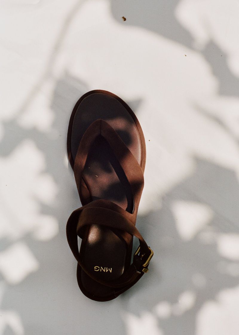 Overhead shot of a single brown Mango brand sandal with a heel, ankle strap, and toe post, set against a white background with leaf shadows.