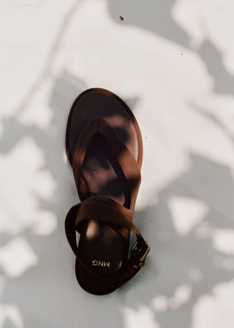 Overhead shot of a single brown Mango brand sandal with a heel, ankle strap, and toe post, set against a white background with leaf shadows.
