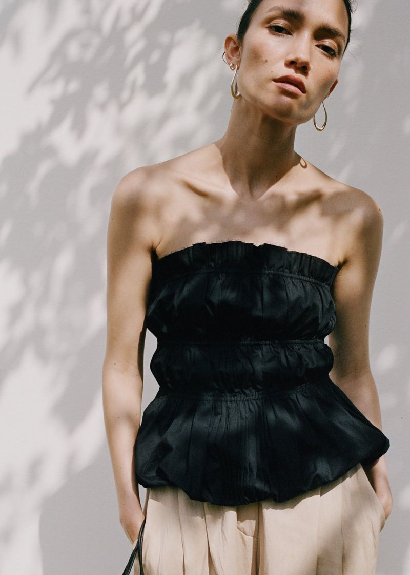 An adult woman poses in natural light wearing a strapless black top and tan pants, with a wall and tree shadows in the background.