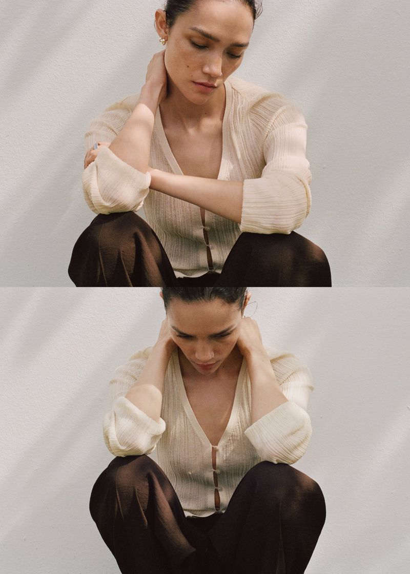 Young woman in a cream blouse and brown pants sits against a white wall with dappled light, looking down in a fashion or editorial shot.