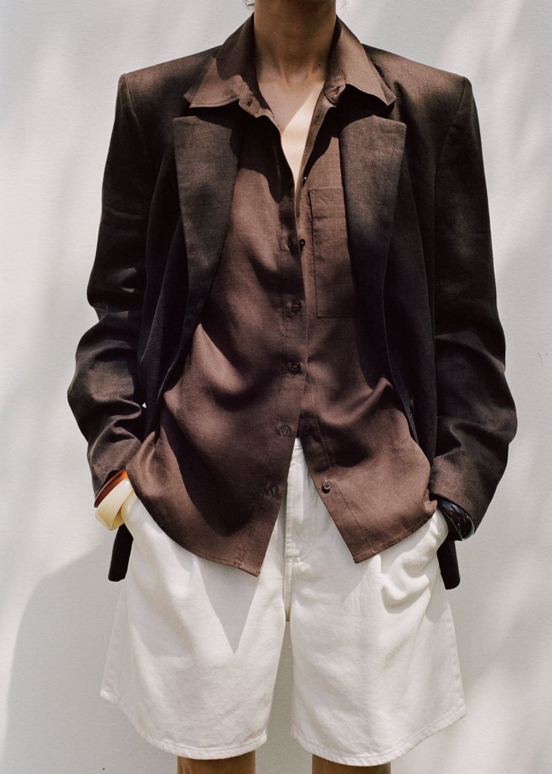 Woman in a dark brown jacket and matching collared shirt, posing in white shorts against a white background with soft shadows.