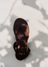 Top-down view of a single brown leather thong sandal with an ankle strap set against dappled light and shadows.