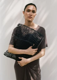 Model Charlotte Carey holding an oversized black woven mesh bag against a pale wall with soft shadows for Mango Urban Summer SS25.
