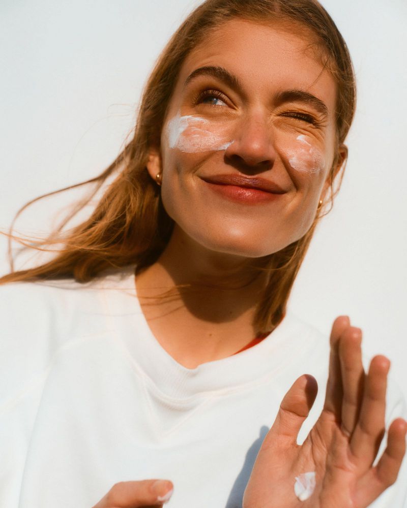 Close-up portrait of a smiling woman with sunscreen cream on her cheeks and hands against a bright white background.