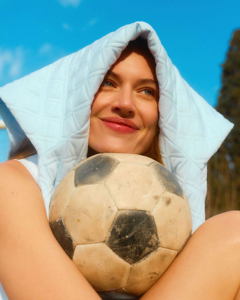 Close-up of a smiling female model holding a worn soccer ball with a light blue quilted cloth draped over her head against a bright sky.