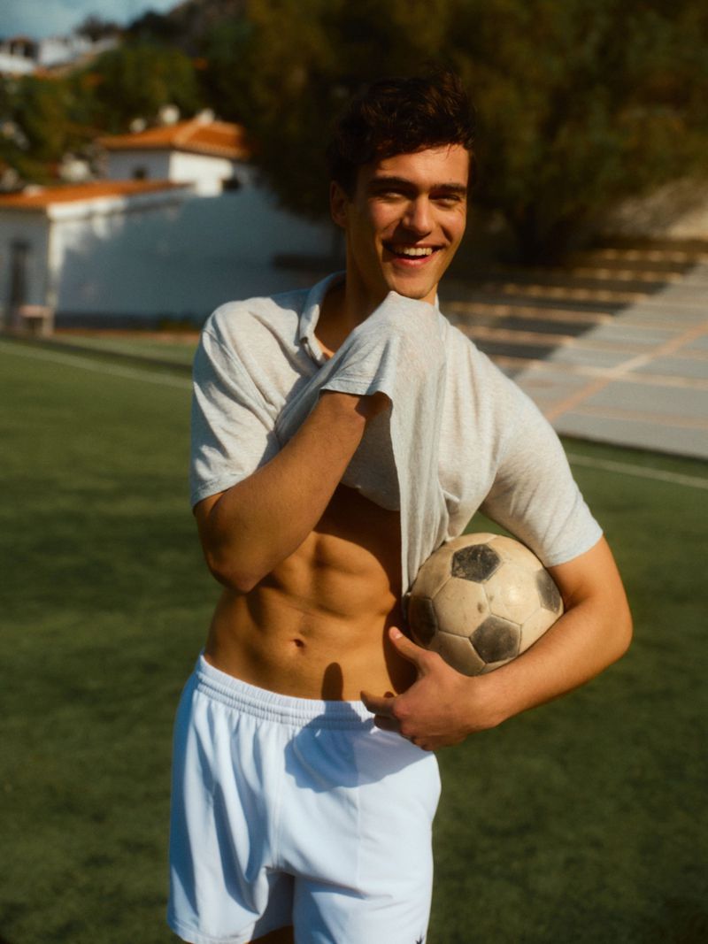 Smiling man holding a soccer ball on a grassy field wearing a white shirt and shorts