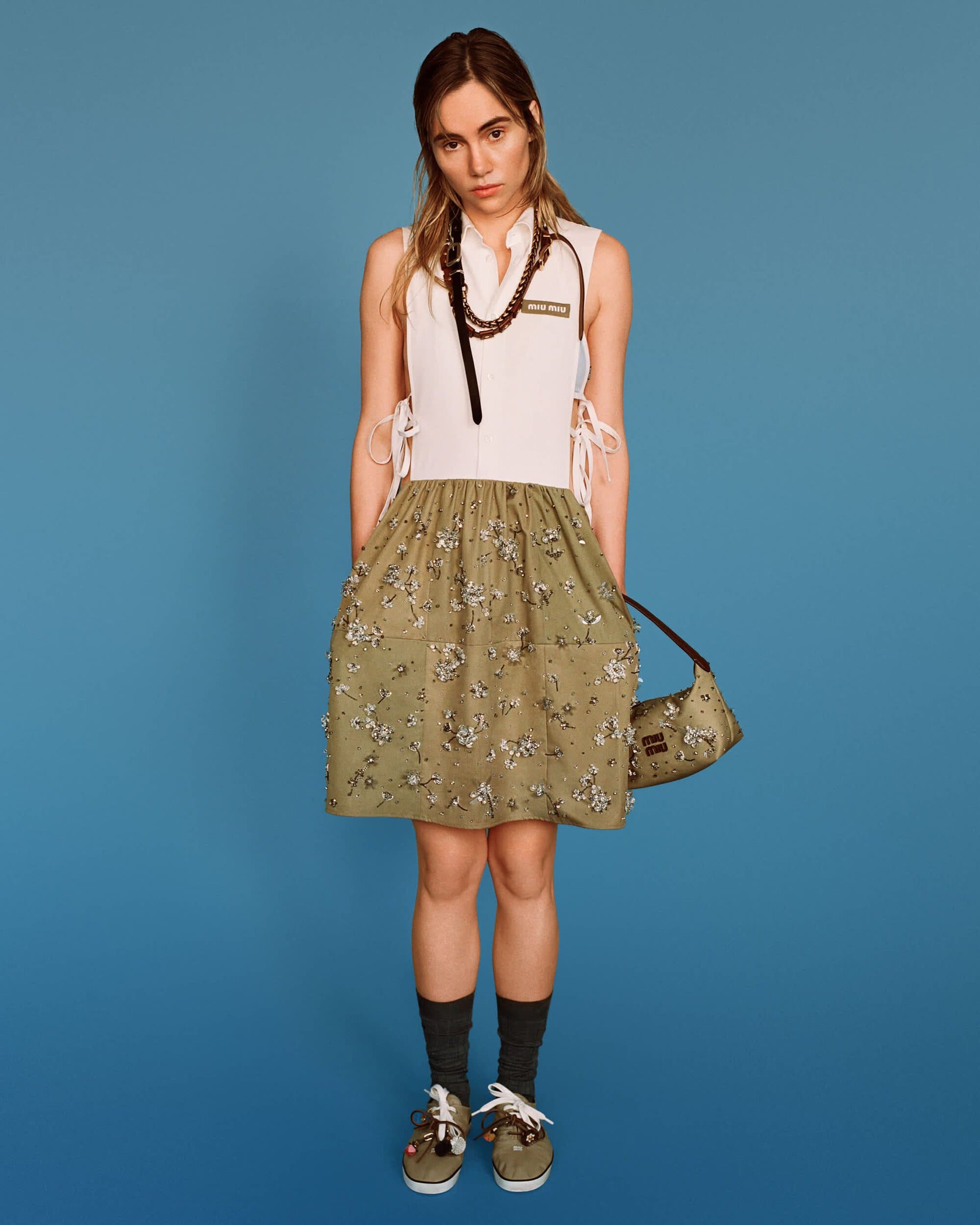 Model wearing a white sleeveless top and olive patterned skirt holding a small handbag against a blue background