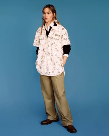 Model standing against a vibrant blue backdrop wearing a patterned white shirt over a black layer and tan trousers.