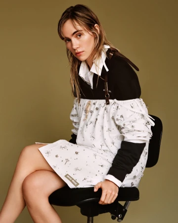 Model seated on a black office chair against an olive green backdrop wearing a white and black patterned dress.