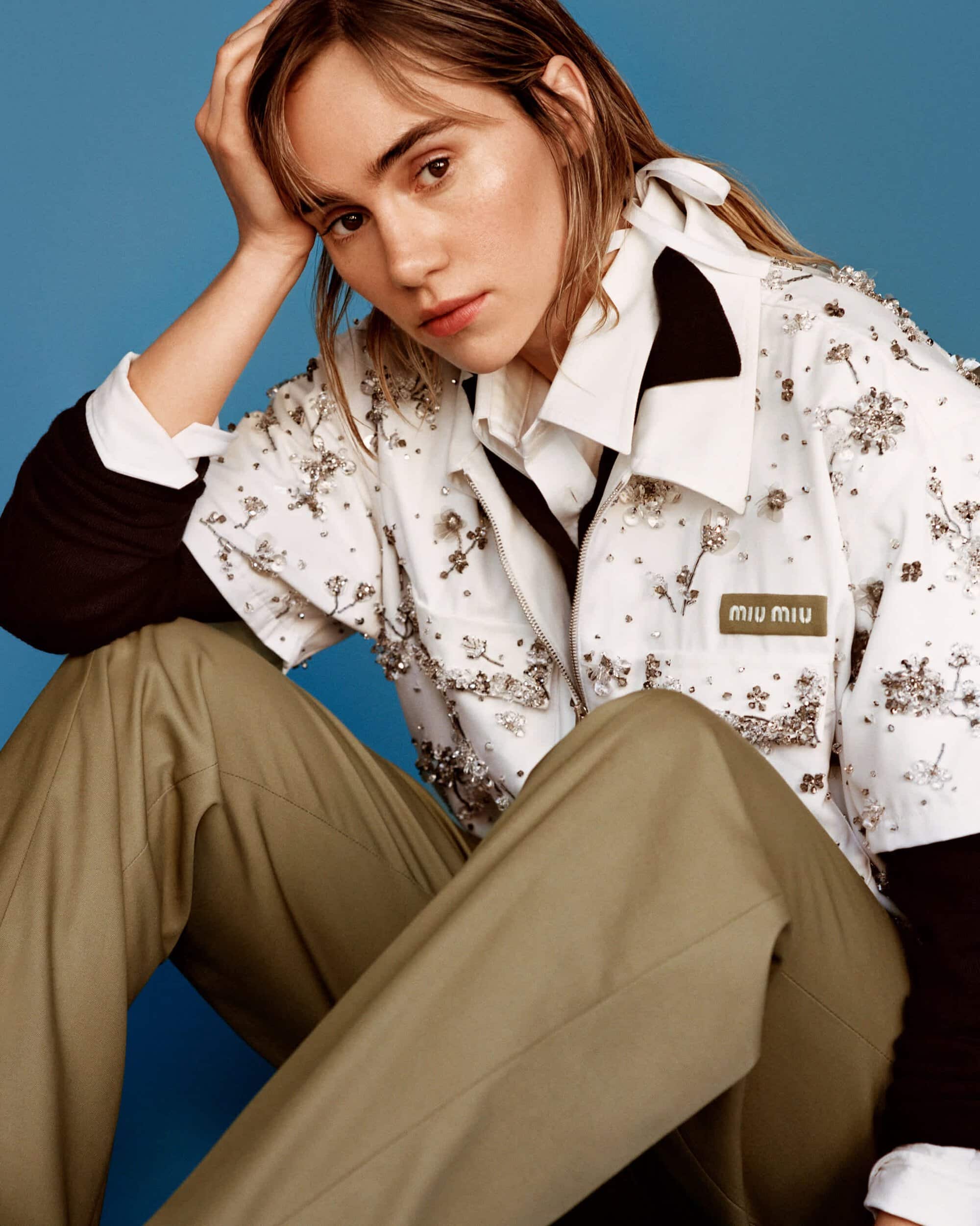 Model seated wearing an embellished white shirt and khaki trousers against a solid blue studio backdrop