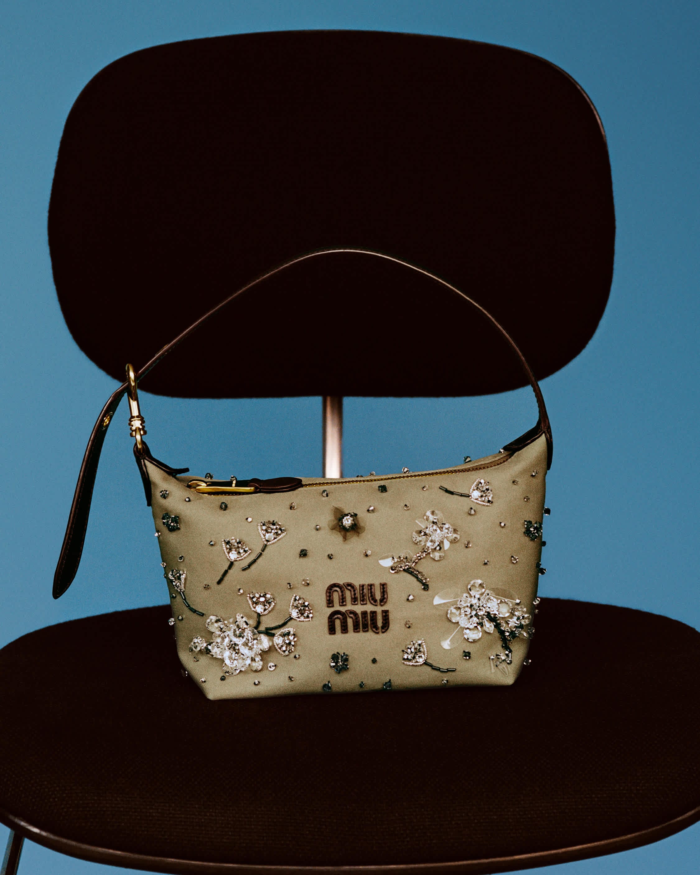 Embellished Miu Miu shoulder bag resting on a dark chair against a solid blue background.