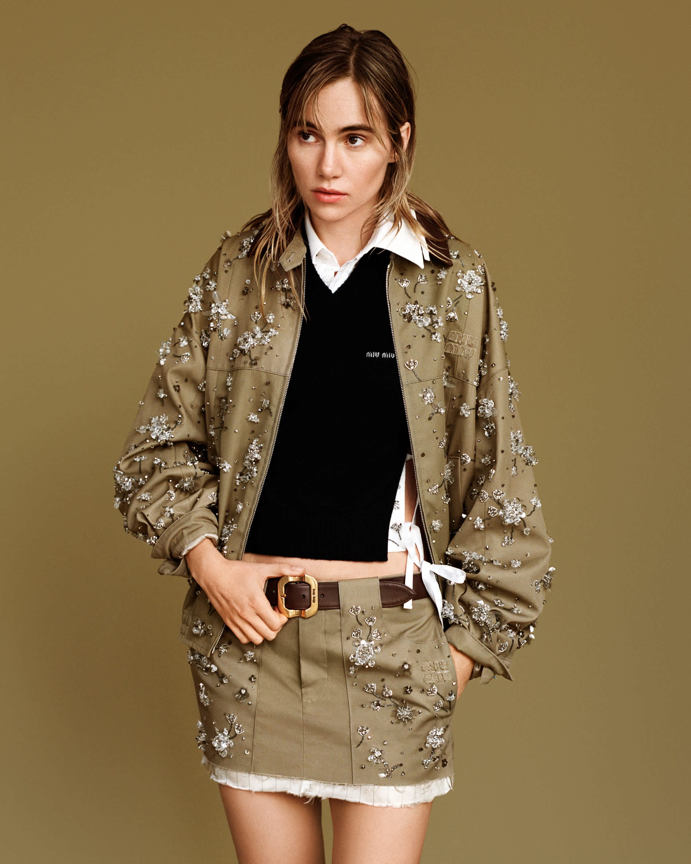 Model wearing an embellished olive jacket and skirt over a black top against a solid olive backdrop