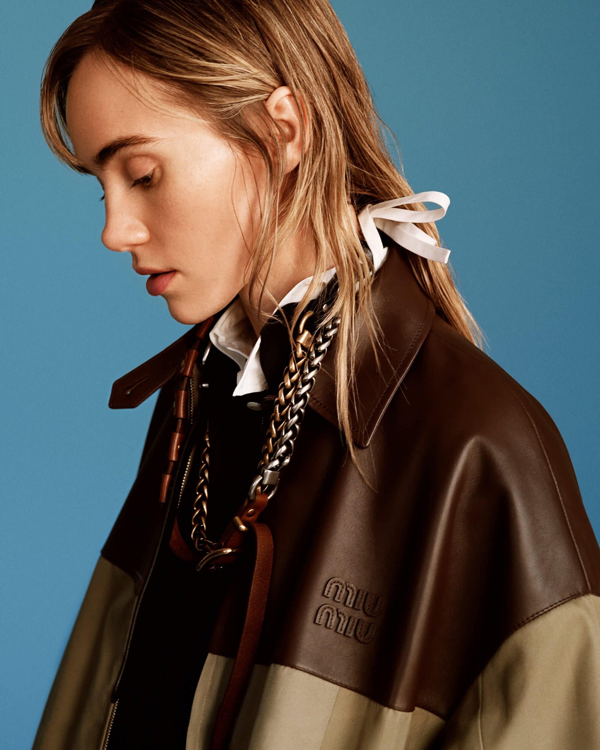 Model in brown and olive jacket with a white collar and ribbon in hair against a solid blue backdrop