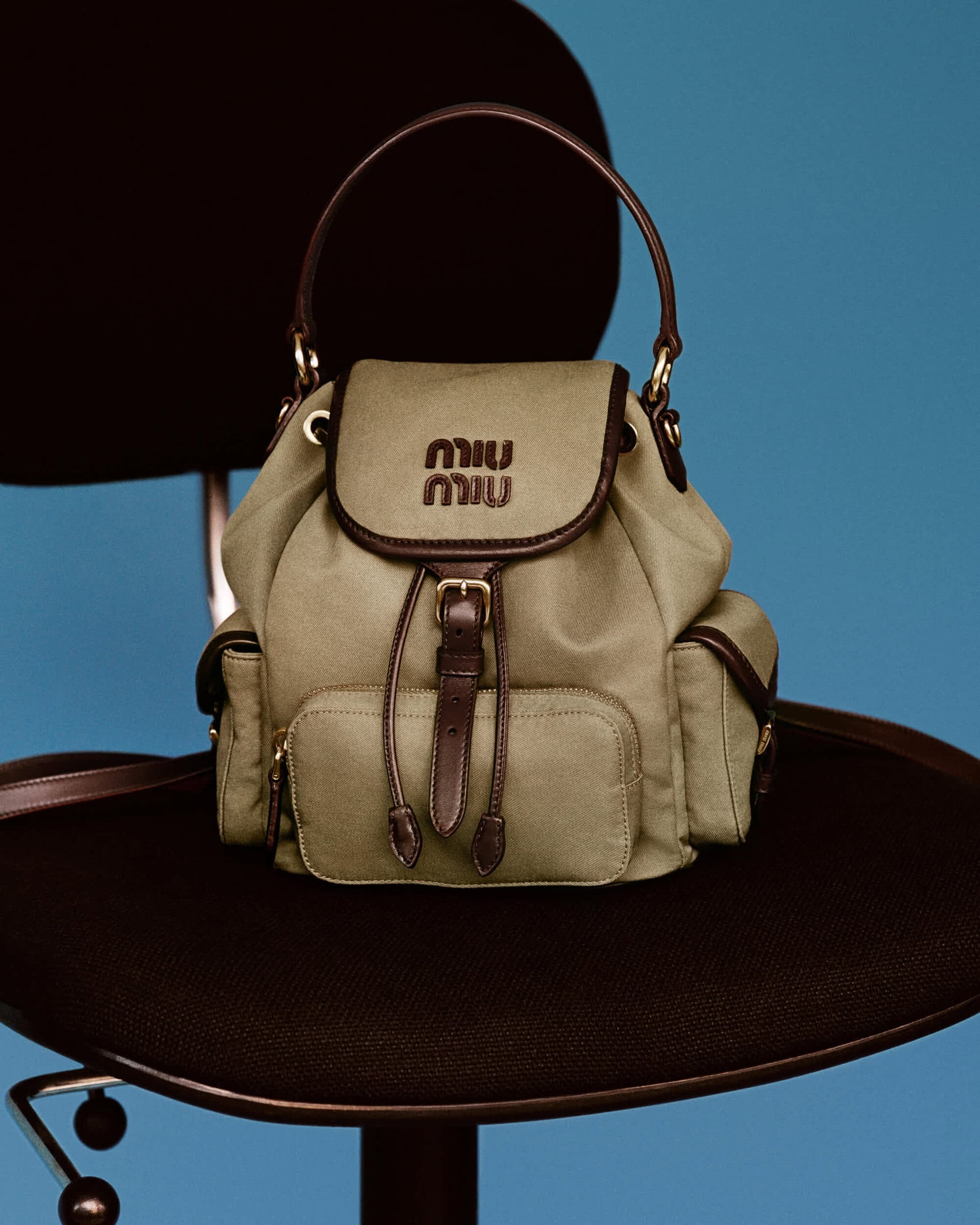 Miu Miu olive green canvas backpack with brown leather trim resting on a black office chair against a blue background