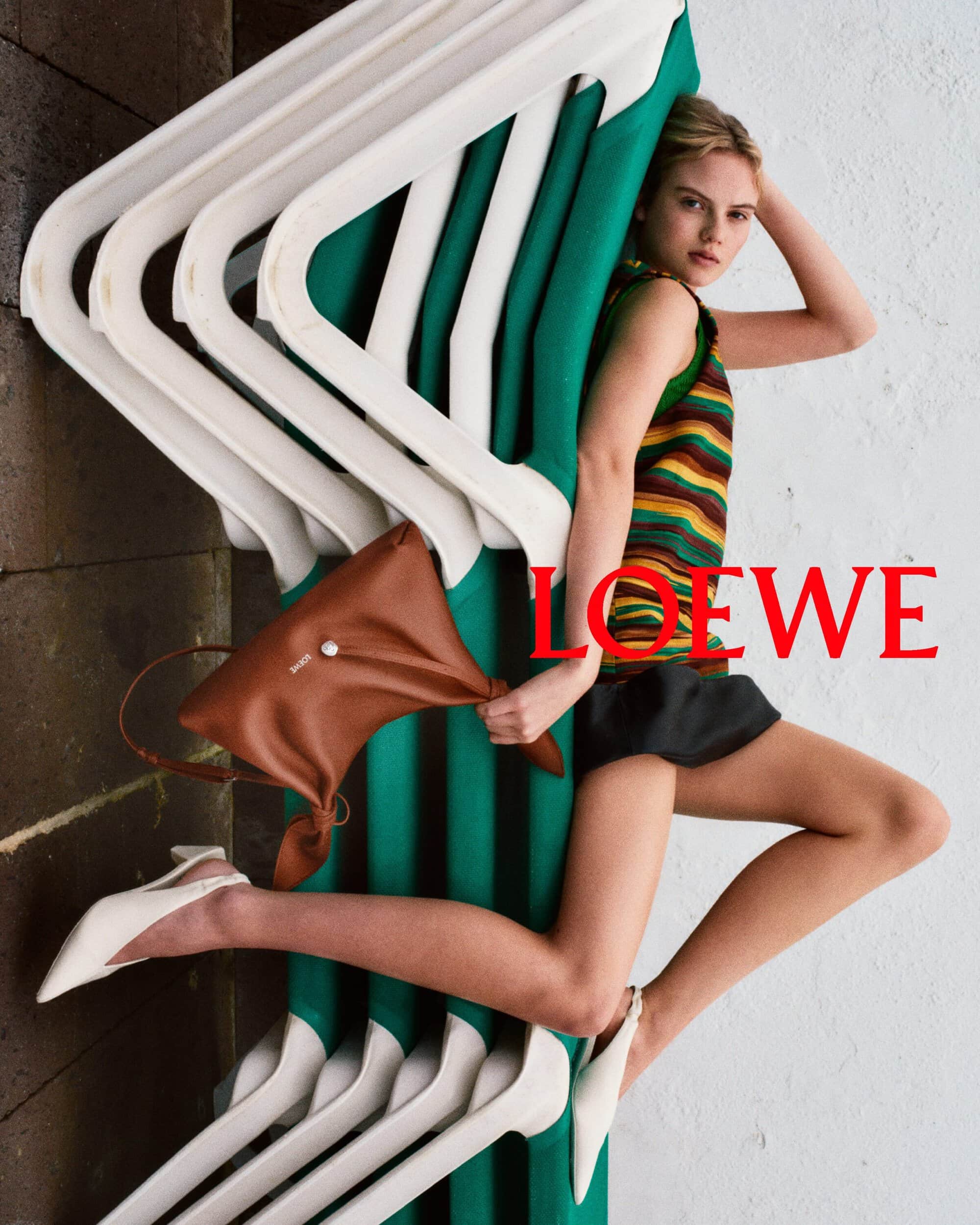 Model leaning against stacked green and white loungers holding a brown leather handbag in a Loewe campaign image