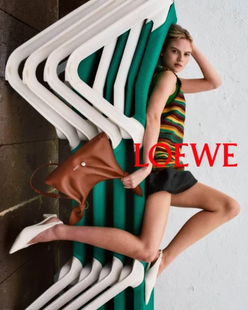 Model leaning against stacked green and white loungers holding a brown leather handbag in a Loewe campaign image