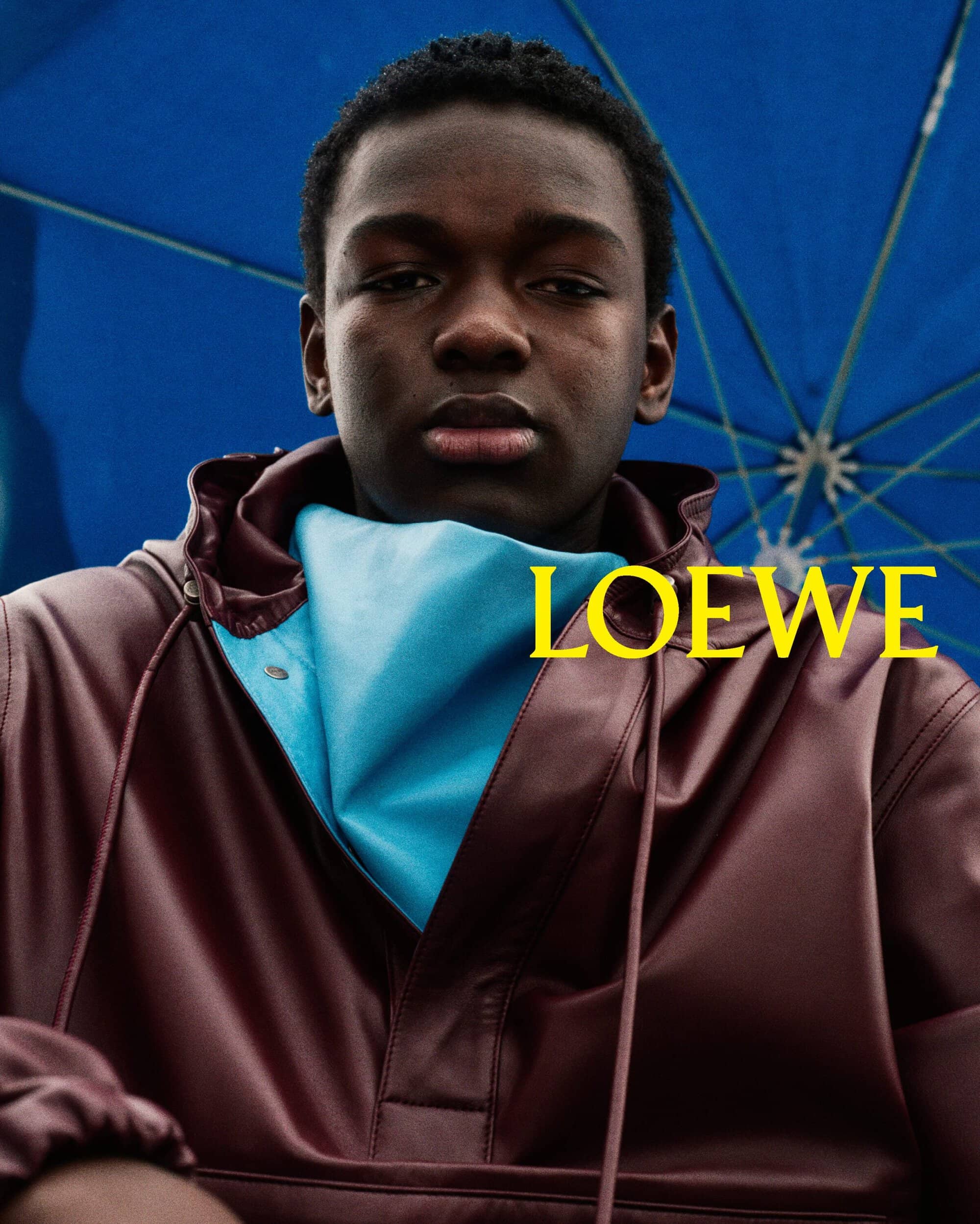 Close-up portrait of a Black male model wearing a dark burgundy leather jacket with a blue neck scarf under a blue umbrella for Loewe